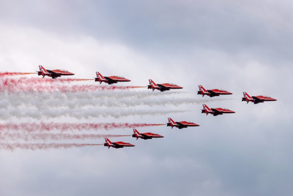 Low angle of modern red military aircraft flying in cloudy sky symmetrically and creating lines with color smokes during air show